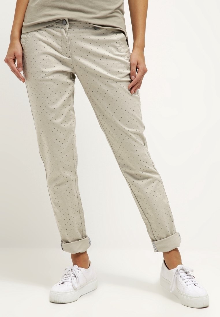 Femme portant un pantalon beige clair ajusté avec de petits pois foncés, ourlets retroussés, et des baskets blanches à lacets, debout sur un sol blanc.
