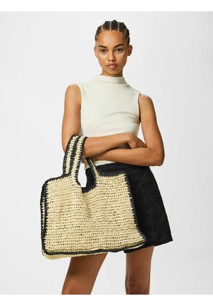 Kvinde med flettet hår bærer ærmeløs creme top og sorte shorts, holder stor vævet beige tote taske med sort kant mod hvid baggrund.
