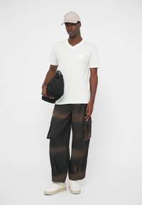 T-shirt branca de manga curta com decote em V e pequeno logotipo no peito; calças cargo pretas com padrões em cor ferrugem; segurando uma bolsa preta; a usar um boné bege.