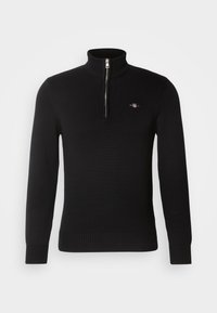 Svart ribbestrikket genser med lange ermer, høy krage og sølvfarget frontglidelås, med en liten brodert logo på venstre bryst.