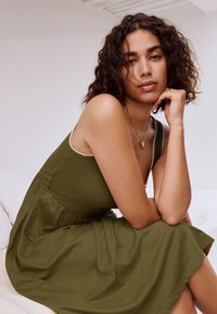 Olivengrøn kjole i ribbet tekstur, tætsiddende overdel med spaghettistropper og en skørt, der flår ud. Synlig guldkæde som accent.