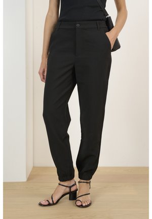 Frau trägt schwarze, hoch taillierte, konisch geschnittene Hosen mit elastischen Bündchen und schwarze, mit Riemchen versehene Sandalen mit Absatz, eine Hand in der Tasche, steht drinnen.