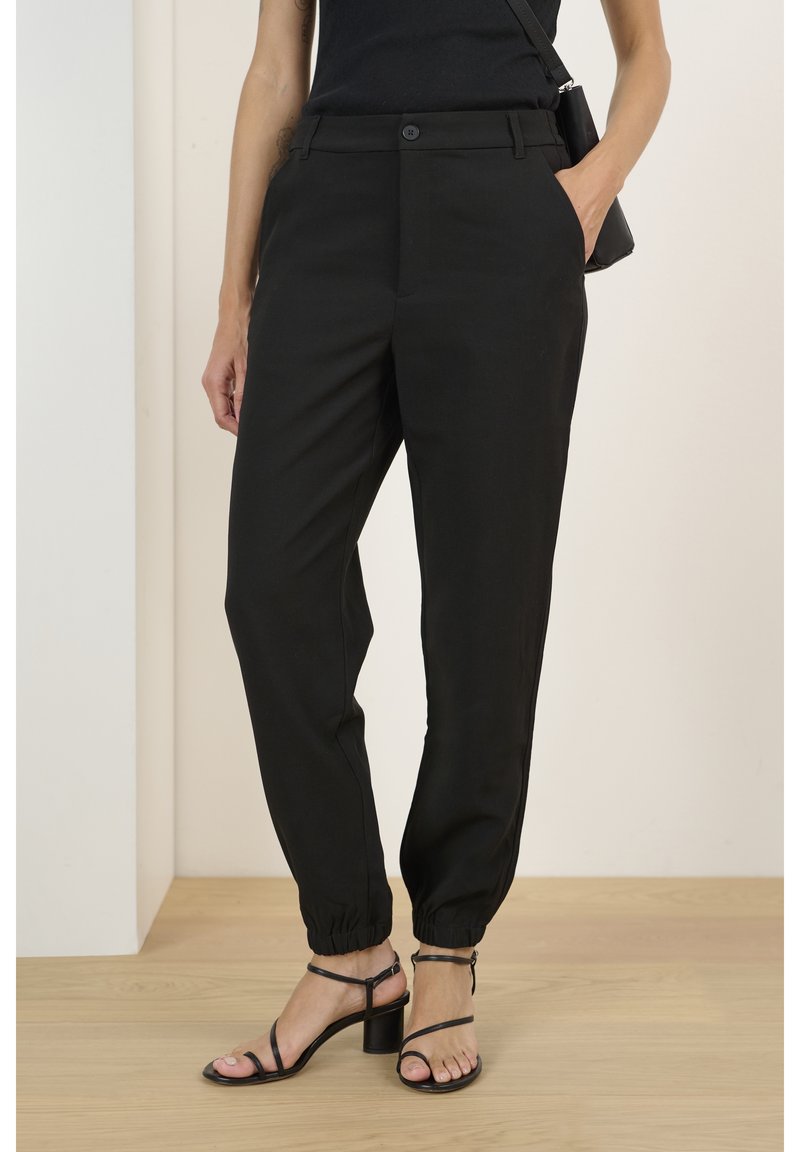 Femme portant un pantalon noir taille haute à jambes fuselées avec des poignets élastiques et des sandales à talons noires à lanières, une main dans la poche, debout à l'intérieur.