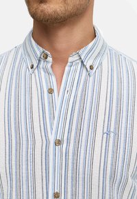 Camisa de botones a rayas en tela ligera, con rayas azules y negras sobre un fondo blanco. Incluye detalles de botones y bordado de logo.