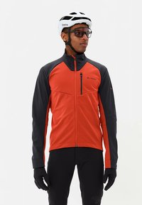 Rood-grijze softshell jas met een hoge kraag, een borstzak met rits en lange mouwen. Draagt zwarte handschoenen en een helm.