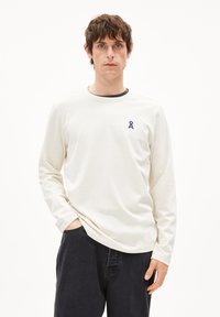Lange mouwen crèmekleurig T-shirt gemaakt van zacht materiaal, met een ronde halslijn en een klein paars lintlogo op de borst.