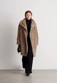 Φουσκωτό μπεζ oversized παλτό με φαρδύ γιακά, συνδυασμένο με μαύρο ζιβάγκο και παντελόνι. Μια μικρή μαύρη τσάντα ώμου προσθέτει λεπτομέρεια.