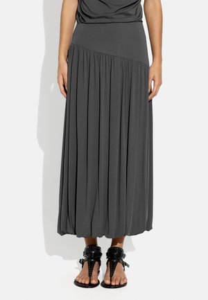 Kobieta w długiej, ciemnoszarej plisowanej spódnicy z ukośnym paskiem w talii i czarnych, płaskich sandałach z ćwiekami na białym tle.