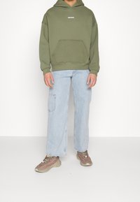 Personne portant un sweat à capuche vert olive avec poche kangourou, un jean large bleu clair avec poche latérale et des baskets beige et rose, debout.