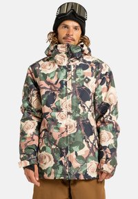 Mężczyzna w kwiecistej kurtce zimowej, czarnej beanie i goglach narciarskich na głowie, stojący na tle białego tła.