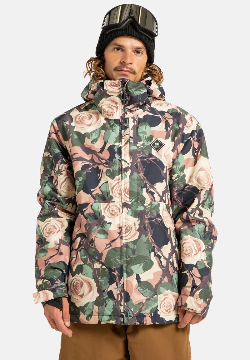 Mężczyzna w kwiecistej kurtce zimowej, czarnej beanie i goglach narciarskich na głowie, stojący na tle białego tła.