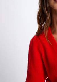 Roter Wollpullover mit V-Ausschnitt und strukturiertem Strickmuster. Die Ärmel sind leicht locker, und der Stoff hat ein weiches, flauschiges Aussehen.