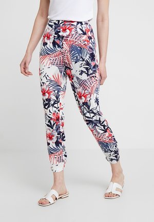 Mujer con sandalias blancas y pantalones con un patrón floral de hojas y flores tropicales rojas, azul marino y rosas sobre un fondo blanco.