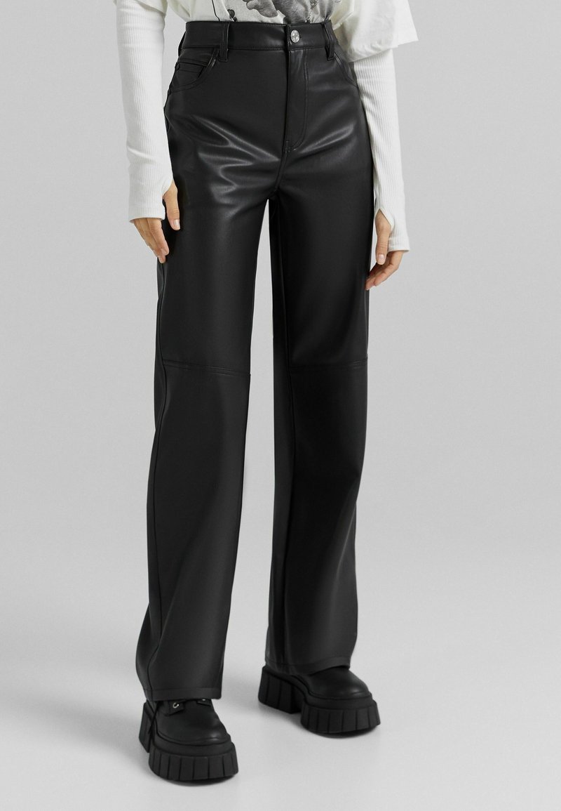 Femme portant un pantalon large en similicuir noir taille haute, des chaussures compensées noires surdimensionnées et un haut blanc à manches longues.