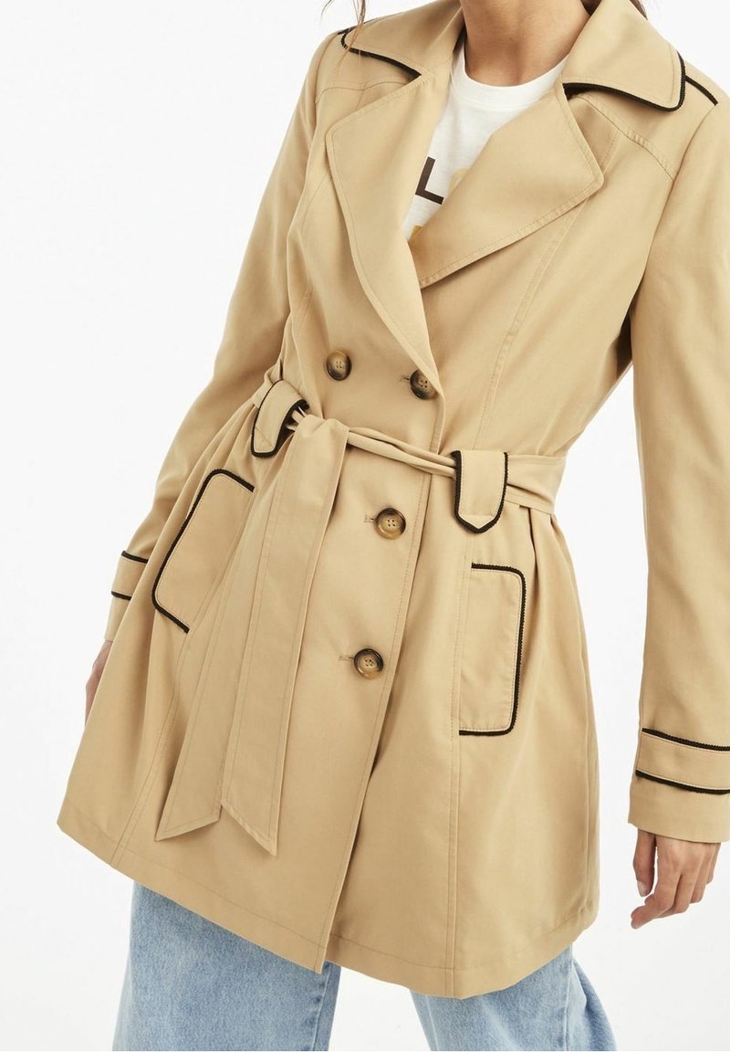 Trench-coat beige croisé avec bordures noires, taille ceinturée et grandes poches, porté sur un jean bleu clair.