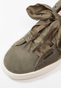 Gros plan sur une basket en daim vert olive avec de larges lacets en ruban de satin noués en nœud et une semelle en caoutchouc blanche sur fond blanc.