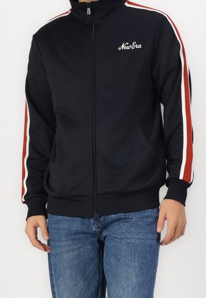 Mand iført en sort lynlåstrøje med røde og hvide striber på ærmerne og New Era-logo, kombineret med blå jeans.