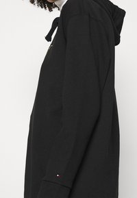 Černá mikina s kapucí a šňůrkami, s logem na hrudi, měkká látka, dlouhé rukávy a kontrastní znak na manžetě.