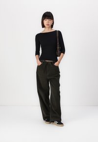 Zwarte, lange mouwen top gecombineerd met wijde olijfgroene corduroy broek; model staat op witte achtergrond met zwart-witte sneakers.