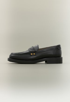 Mocasín de cuero negro para hombre con costura de puntera mocasín, tira decorativa con dos tachuelas doradas y tacón bajo apilado sobre fondo neutro.