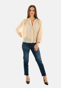 Lichtbeige, semi-doorzichtige blouse met een knoopsluiting aan de voorkant en geplooide details op de schouders, gecombineerd met donkerblauwe skinny jeans en zwarte hakken.