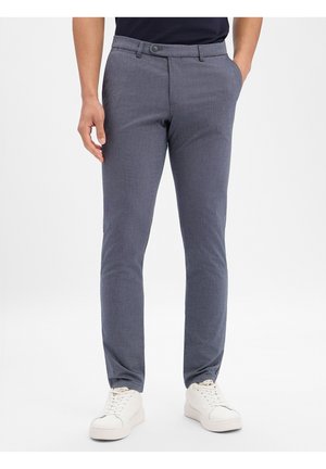 Mann trägt enganliegende graue Hosen, weiße Sneakers und ein marineblaues Hemd, steht mit einer Hand in der Tasche vor einem schlichten Hintergrund.