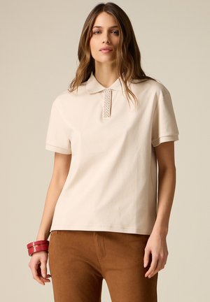 Vrouw draagt een beige polo met korte mouwen versierd met kralen bij de kraag, bruine broek en twee rode armbanden om haar linkerpols.