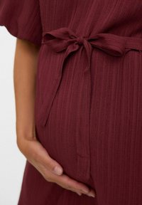 Vestido de tela texturizada color burdeos con cintura atada, con rayas verticales y mangas cortas, que se ajusta suavemente sobre un vientre redondeado.