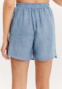 Světle modré lněné šortky s elastickým pasem, volného střihu a bočními rozparky pro snadný pohyb. Bez viditelných kapes.
