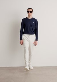 Camisa de manga larga azul marino, pantalones joggers blancos con parche rojo, y zapatillas blancas. Gafas de sol negras completan el conjunto. Fondo neutro.