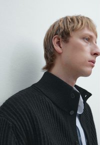 Black ribbed knit sweater with a high collar and button closure, worn over a light striped shirt. Smooth texture with subtle detailing.
