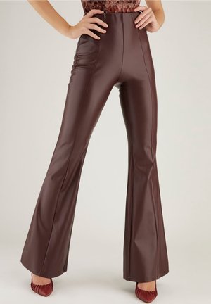 Femme portant un pantalon évasé en cuir marron foncé taille haute et des talons rouges à bout pointu, les mains posées sur les hanches sur un fond uni.