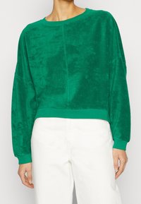 Zelená zkrácená mikina vyrobená z měkké, texturované tkaniny. Má dlouhé rukávy a kulatý výstřih, s jednolitou barvou po celém povrchu.