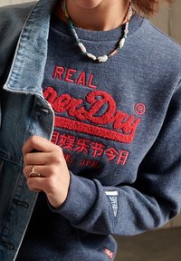 Sudadera azul marino con el logotipo bordado en rojo "Superdry". Mangas largas, cuello redondo y un parche con "Superstate" en el puño. Chaqueta de mezclilla parcialmente visible.