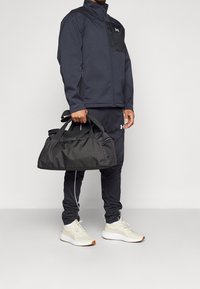 Svart zip-jacka med grå detaljer, i kombination med en matchande svart duffelväska. Bärs med svarta joggers och ljusa sneakers.