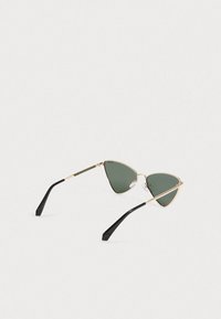 Sončne očala v obliki mačjih oči z zlatimi kovinskimi okvirji, temno zelenimi lečami in črnimi ukrivljenimi ročaji, ki ležijo na beli površini.