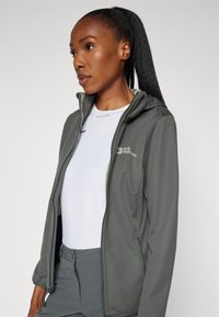 Grijze hoodie gemaakt van zachte, rekbare stof, voorzien van een ritssluiting aan de voorkant en een logo op de linkerborst. Draagt een wit shirt eronder.