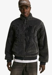 Black fuzzy jacket with a zip closure, featuring a textured design and ribbed cuffs, layered over a white shirt. Dark jeans complement the look.