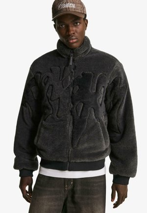 Črna puhasta jakna z zadrgo, s teksturiranim dizajnom in rebrastimi manšetami, slojeno čez belo majico. Temne kavbojke dopolnjujejo videz.