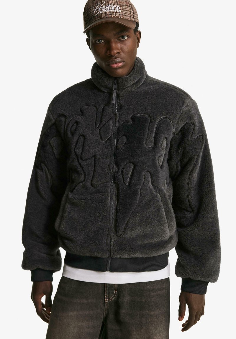 Black fuzzy jacket with a zip closure, featuring a textured design and ribbed cuffs, layered over a white shirt. Dark jeans complement the look.