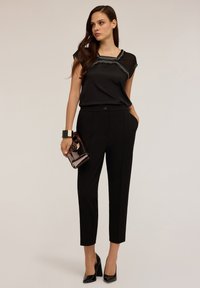 Zwarte blouse met doorschijnende mouwen en versierde halslijn, gecombineerd met ge-tailored zwarte broek en glanzende zwarte hakken, met een clutch in de hand.