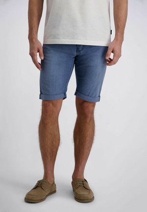 Mann trägt hochgekrempelte blaue Jeansshorts, weißes T-Shirt und braune Schnürschuhe, steht vor einem einfarbigen Hintergrund.