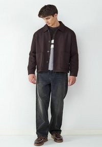 Ciemnobrązowa kurtka z kołnierzykiem, dwoma przednimi kieszeniami i guzikami; noszona na szarym swetrze w paski oraz zestawiona z luźnymi dżinsami.