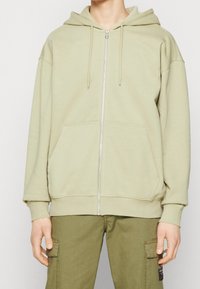 Sudadera con cremallera de color verde claro, con un bolsillo frontal, capucha con cordón y puños acanalados. Hecha de un tejido suave, presenta un ajuste relajado.