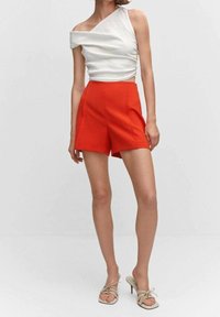 Kvinne iført en asymmetrisk hvit ermeløs topp, høyt midje rød shorts og beige åpne hælsandaler som står foran en ensfarget bakgrunn.