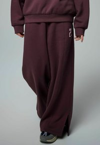 Mörk röd oversized sweatpants med sidofickor, tillverkade av mjukt tyg. Har ett litet rosa nummer "21" på framsidan, kombinerat med grå sneakers.