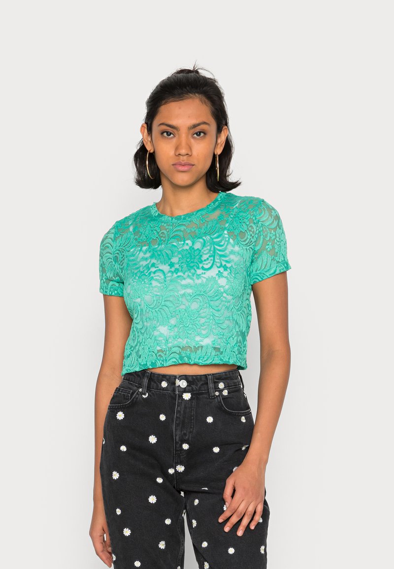 Top curto de renda teal com mangas curtas apresenta padrões florais. Combinado com jeans pretos de cintura alta adornados com motivos de margaridas brancas.