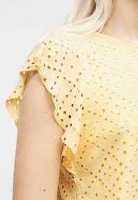 Haut en tissu à œillets jaune avec manches volantées porté par une personne à la peau claire et aux cheveux blonds, cadré au niveau des épaules et du cou.