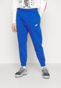 Siniset collegehousut, joissa on joustava vyötärönauha ja resorit nilkoissa, vasemmassa reidessä valkoinen Nike-logo ja sileä, pehmeä pinta.