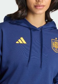 Marineblauer Kapuzenpullover mit gelbem Adidas-Logo und Emblem der spanischen Nationalmannschaft. Verfügt über eine Kordelzug-Verschluss und eine weiche Stoffstruktur.